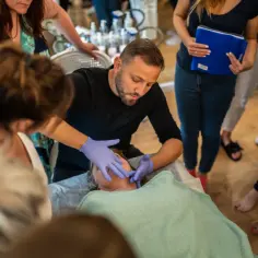 Mężczyzna w rękawiczkach wykonuje masaż liftingujący twarzy. Grupa osób obserwuje, niektórzy z notatkami.