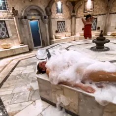 Kobieta w pianie podczas rytuału hammam. W tle inna kobieta w czerwonym ręczniku.