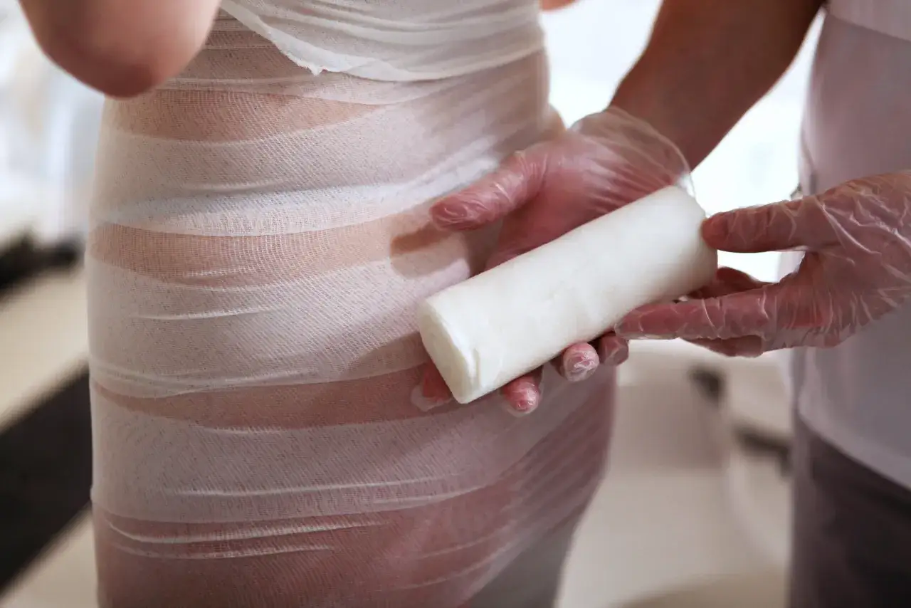 Dłonie w rękawiczkach trzymają rolkę bandaża, przygotowując się do zabiegu body wrapping. Widoczne efekty zabiegu to smuklejsza sylwetka.